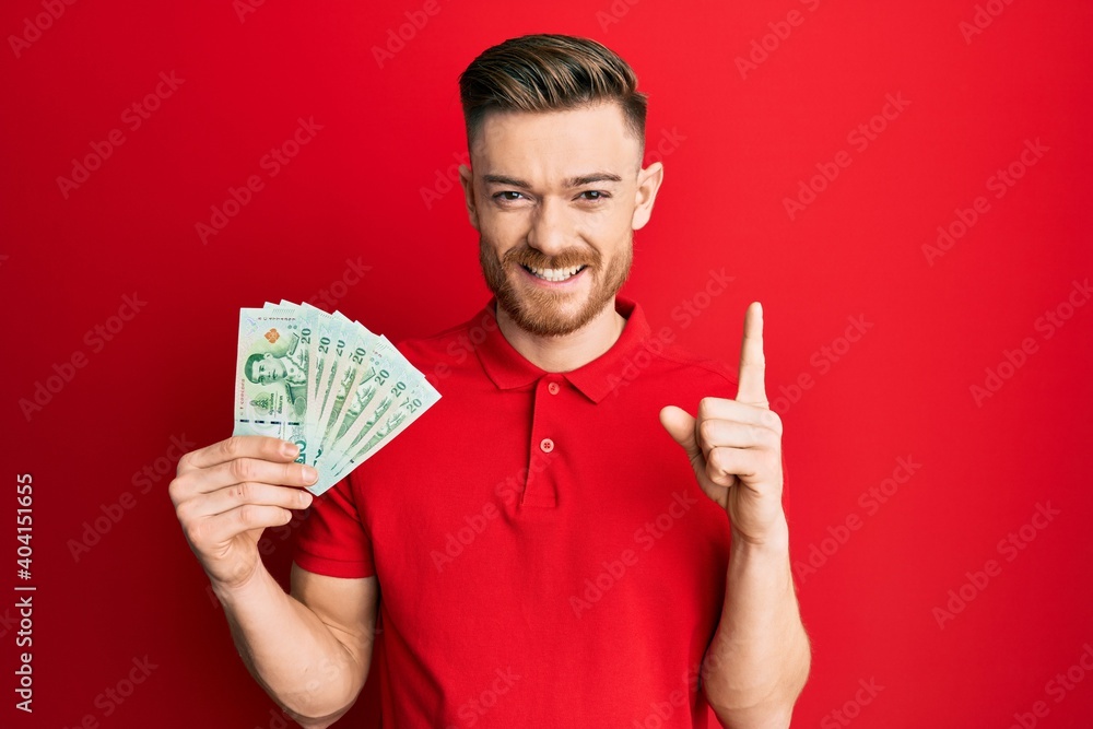 Young redhead man holding 20 thai baht banknotes smiling with an idea or question pointing finger with happy face, number one Young redhead man holding 20 thai baht banknotes smiling with an idea or question pointing finger with happy face, number one