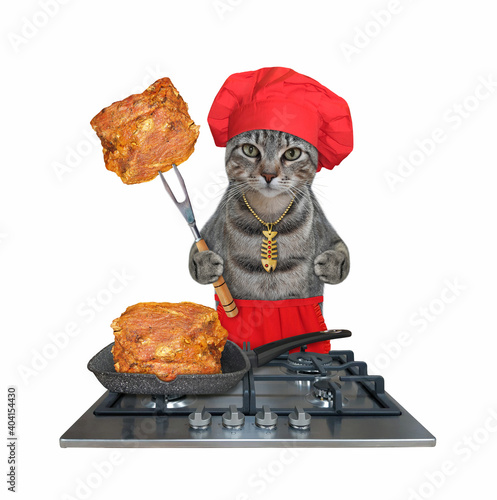 A gray cat in a red chef hat and an apron is cooking fried meat on a gas stove. White background. Isolated.