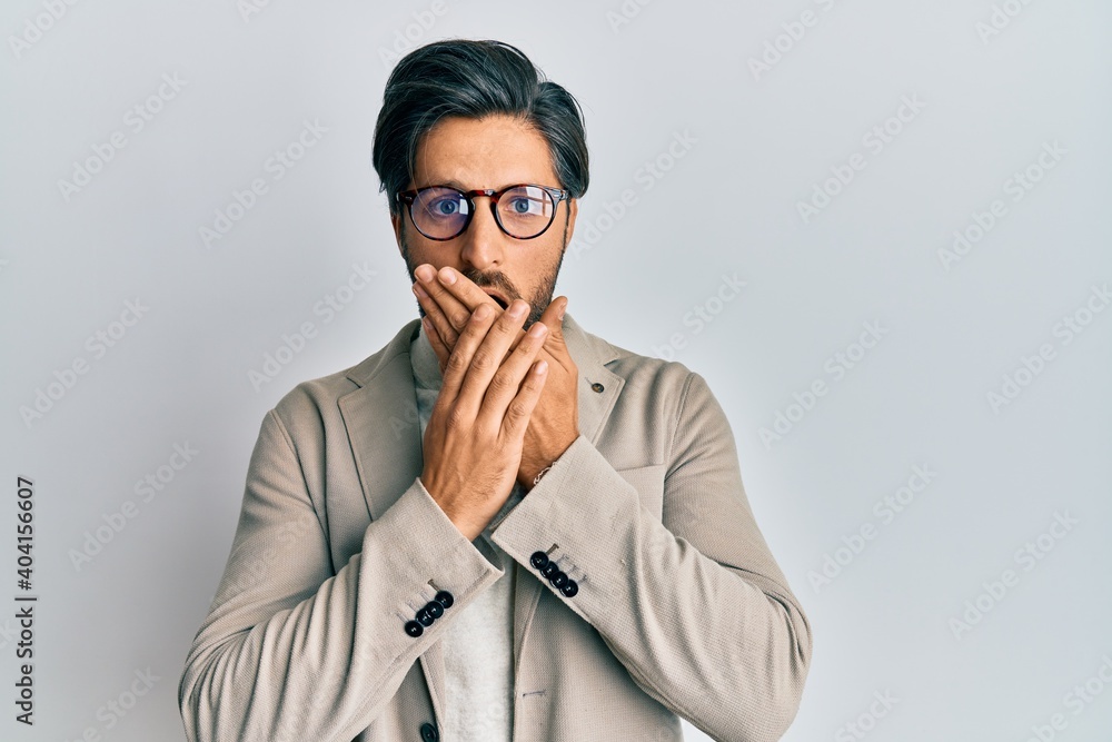 Young hispanic man wearing business jacket and glasses shocked covering mouth with hands for mistake. secret concept. Young hispanic man wearing business jacket and glasses shocked covering mouth with hands for mistake. secret concept.