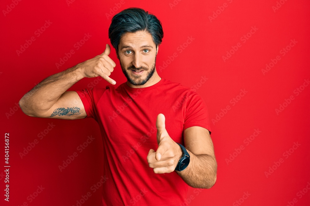 Young hispanic man wearing casual red t shirt smiling doing talking on the telephone gesture and pointing to you. call me. Young hispanic man wearing casual red t shirt smiling doing talking on the telephone gesture and pointing to you. call me.