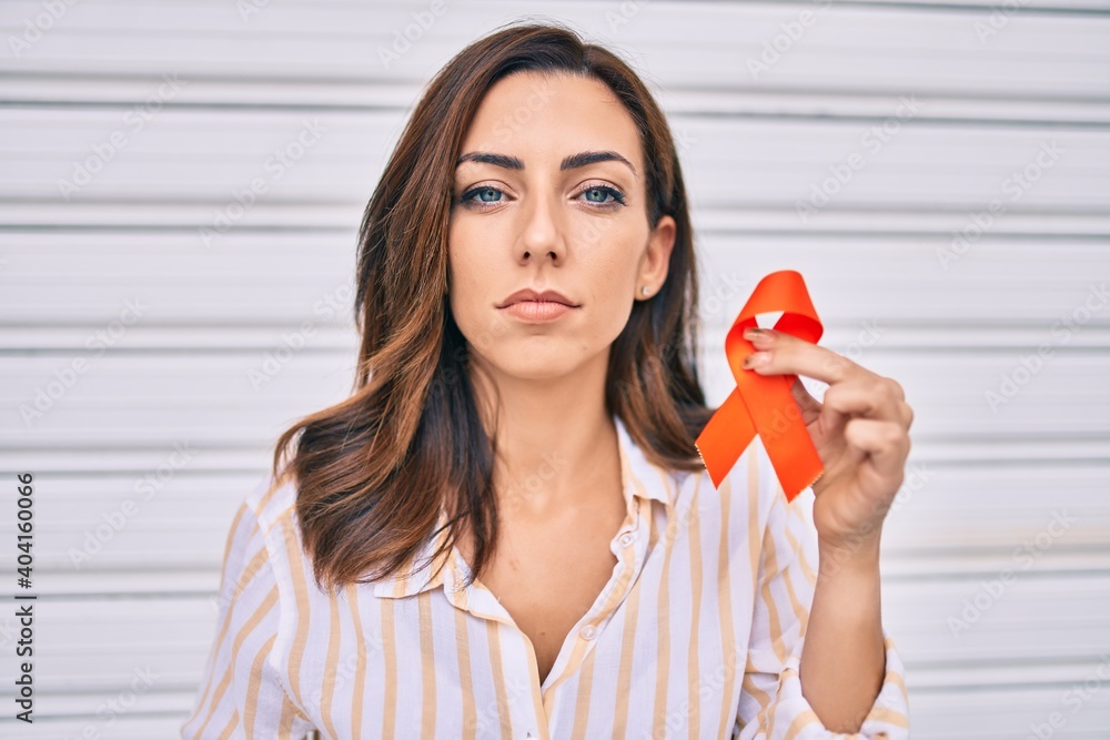 Young hispanic woman with serious expression holding awareness orange ribbon standing at the city. Young hispanic woman with serious expression holding awareness orange ribbon standing at the city.
