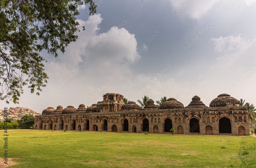 Hampi, Karnataka, India - November 5, 2013: Zanana Enclosure. Brown stone Elephant stables with multiple domes seen over green lawn under blue cloudscape. Green foliage and mountain on horizon.