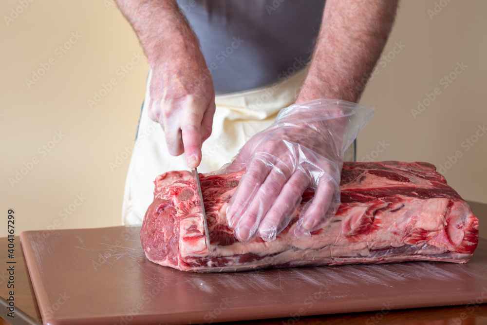 A male butcher or chef is preparing prime rib steaks from a large beef rib roast. The meat is on a white plastic cutting board which is sitting on a restaurant counter. The butcher has a long knife. 