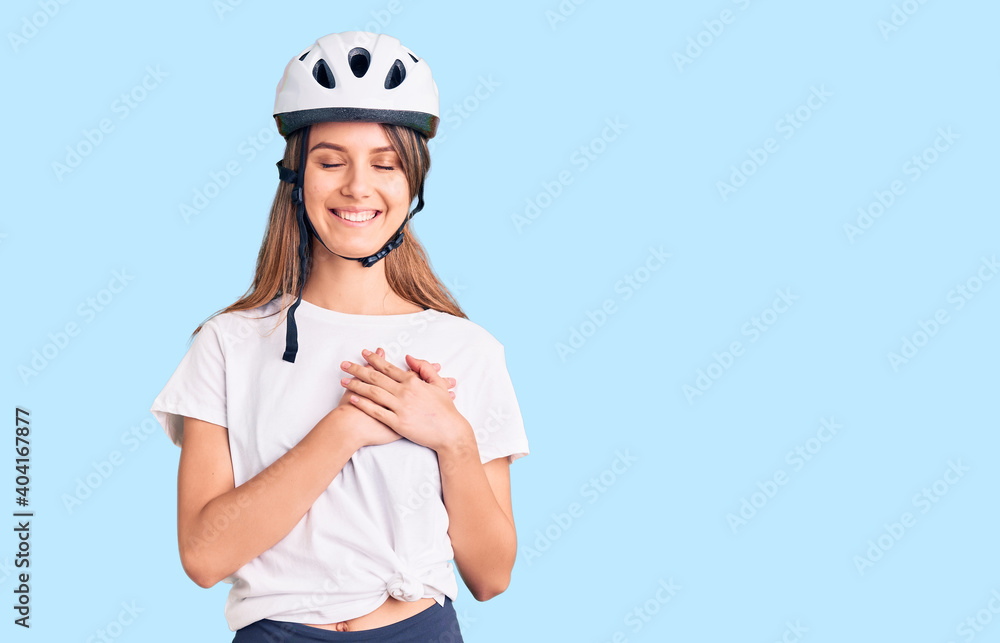 Young beautiful girl wearing bike helmet smiling with hands on chest with closed eyes and grateful gesture on face. health concept. Young beautiful girl wearing bike helmet smiling with hands on chest with closed eyes and grateful gesture on face. health concept.