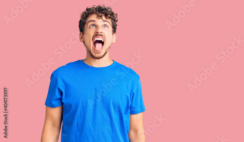 Young handsome man with curly hair wearing casual clothes angry and mad screaming frustrated and furious, shouting with anger. rage and aggressive concept. © Krakenimages.com