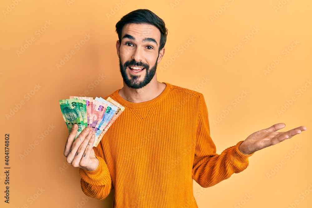 Young hispanic man holding south african rand banknotes celebrating achievement with happy smile and winner expression with raised hand Young hispanic man holding south african rand banknotes celebrating achievement with happy smile and winner expression with raised hand