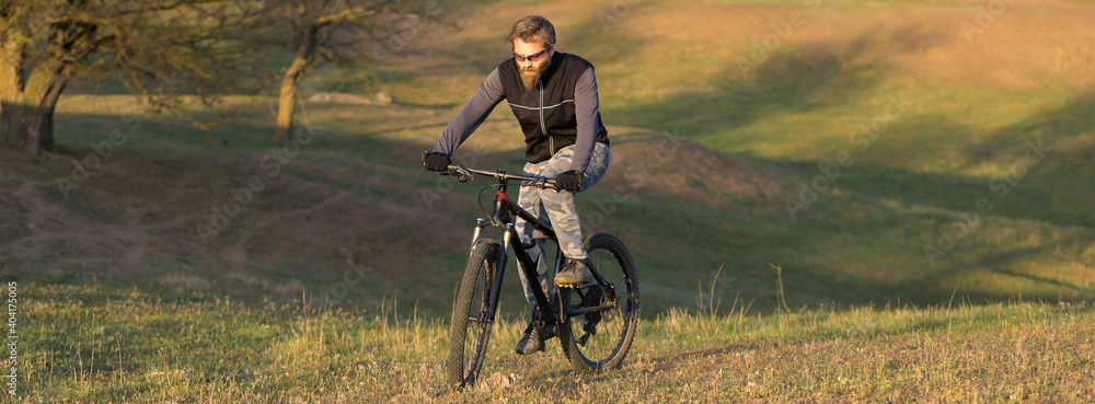 Fototapeta premium Sports brutal bearded guy on a modern mountain bike. Cyclist on the green hills in the spring.