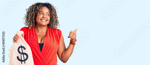 Young african american plus size woman holding money bag with dollar symbol pointing thumb up to the side smiling happy with open mouth