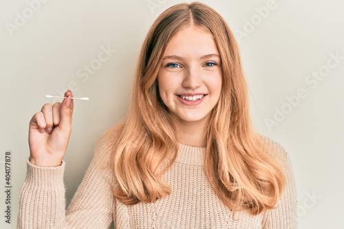 Beautiful young caucasian girl holding earwax cotton remover looking positive and happy standing and smiling with a confident smile showing teeth