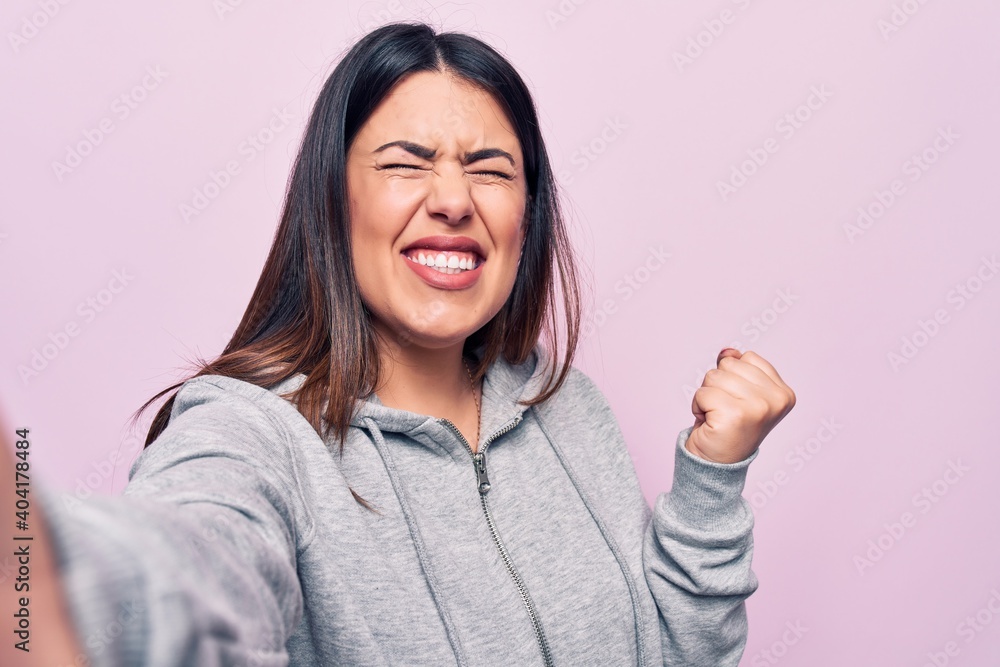 Young beautiful brunette sporty woman make selfie by the camera over pink background screaming proud, celebrating victory and success very excited with raised arm Young beautiful brunette sporty woman make selfie by the camera over pink background screaming proud, celebrating victory and success very excited with raised arm