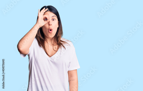 Young beautiful brunette woman wearing casual t-shirt doing ok gesture shocked with surprised face, eye looking through fingers. unbelieving expression.