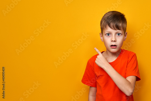 Surprised with open mouth boy in orange t-shirt on yellow isolated background points his finger to the side to the place for text, banner
