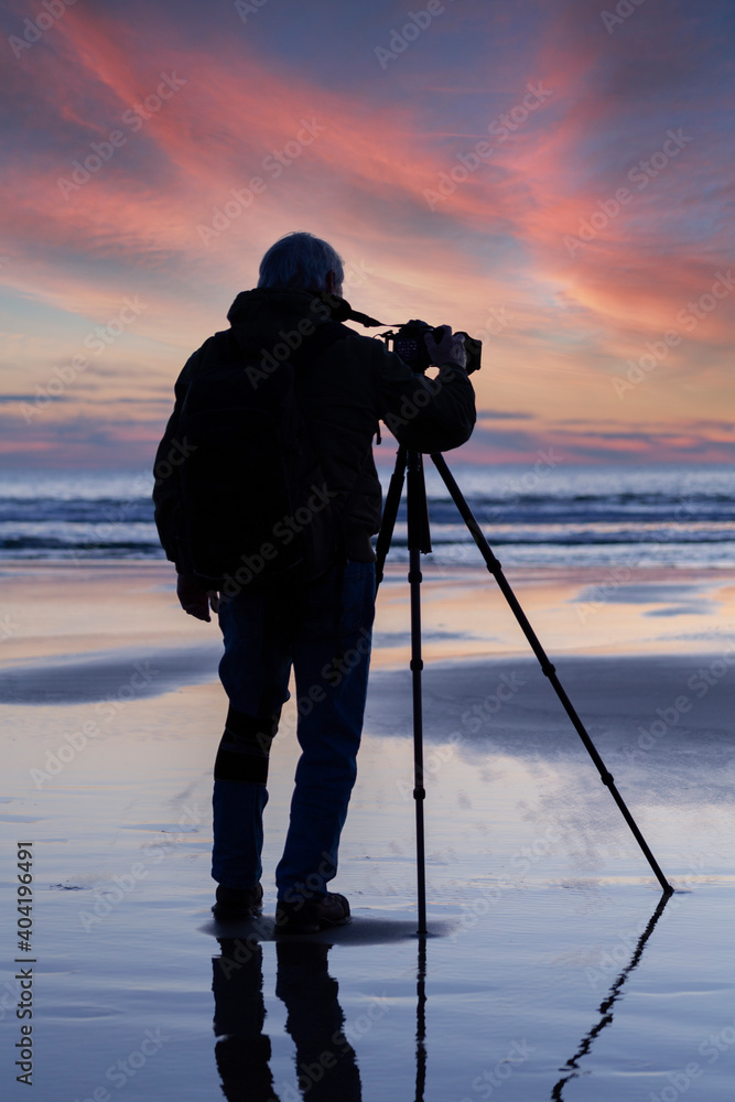 The silhouette of a photographer with a camera on tripod photographing a colorful ocean side sunset