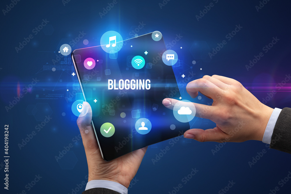 Businessman holding a foldable smartphone with BLOGGING inscription, social media concept Businessman holding a foldable smartphone with BLOGGING inscription, social media concept