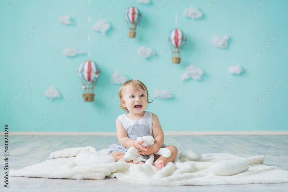 A little Caucasian girl is sitting on the floor against the background of a blue wall decorated with clouds and balloons. Children's room decor.