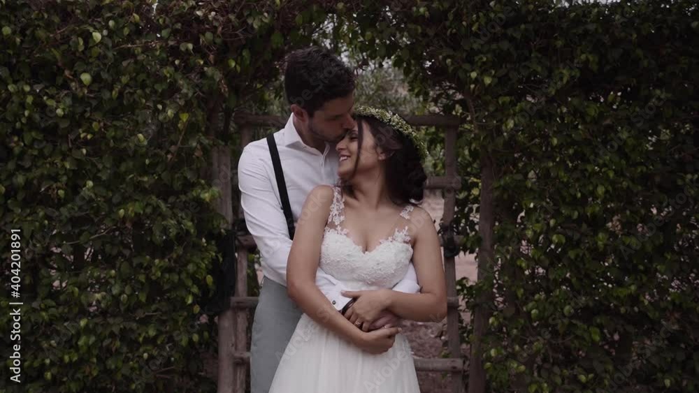 Woman into wonderful white wedding dress looking to her husband who is kissing her forehead standing in front of a green hedge. Dolly shot Woman into wonderful white wedding dress looking to her husband who is kissing her forehead standing in front of a green hedge. Dolly shot