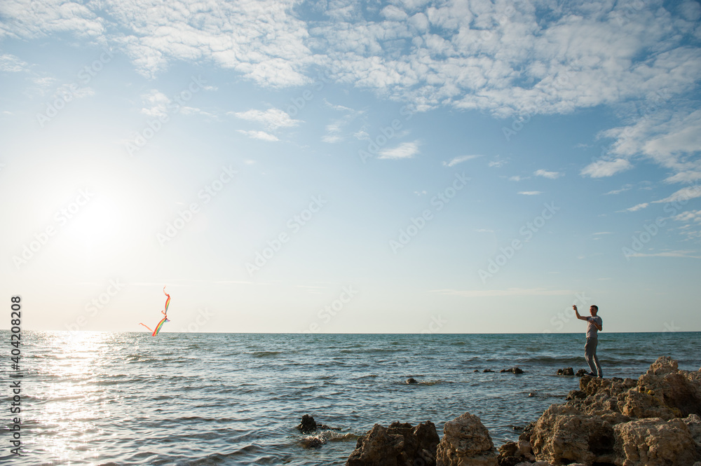 Fototapeta premium one active male playing with flying kite standing on sea rocky coast on sunset summer leisure activity