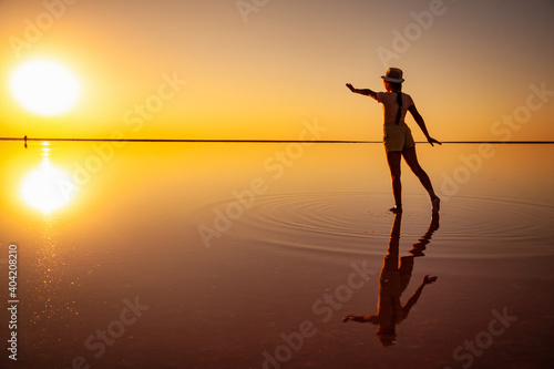 Lovely happy young girl walks and poses in a magical pose along the mirror pink salt lake enjoying the warm evening sun, looking at the fiery sunset and her reflection