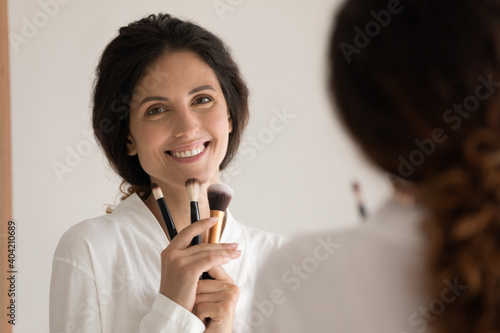 Portrait of smiling young woman look in mirror in home bathroom do daily makeup get ready in morning. Happy millennial Caucasian 20s lady use eco tools brushes for make up. Beauty product concept.