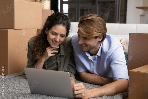 Happy millennial Caucasian man and woman lying on floor in new home on relocation day browsing wireless internet on laptop. Smiling young couple renters search for house rent on computer online.
