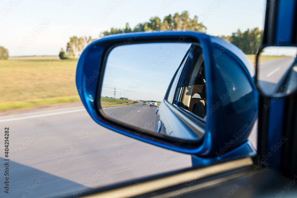 Rear view in the side mirror of a blue sedan with the reflection of a cars on an asphalt road on a summer day with green trees on the sides of the highway.