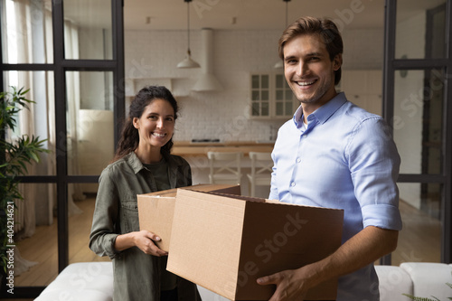 Portrait of smiling young Caucasian man and woman tenants hold cardboard boxes feel excited relocate to new home together. Happy millennial couple first time buyers enjoy moving to own flat or house.