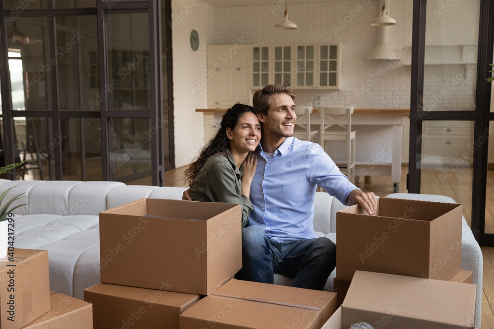 Smiling young Caucasian man and woman sit on sofa hugging look in distance dream together. Happy millennial couple renters enjoy moving relocation day to new own home or house. Rent, estate concept. Smiling young Caucasian man and woman sit on sofa hugging look in distance dream together. Happy millennial couple renters enjoy moving relocation day to new own home or house. Rent, estate concept.