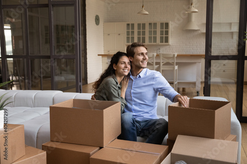 Smiling young Caucasian man and woman sit on sofa hugging look in distance dream together. Happy millennial couple renters enjoy moving relocation day to new own home or house. Rent, estate concept.