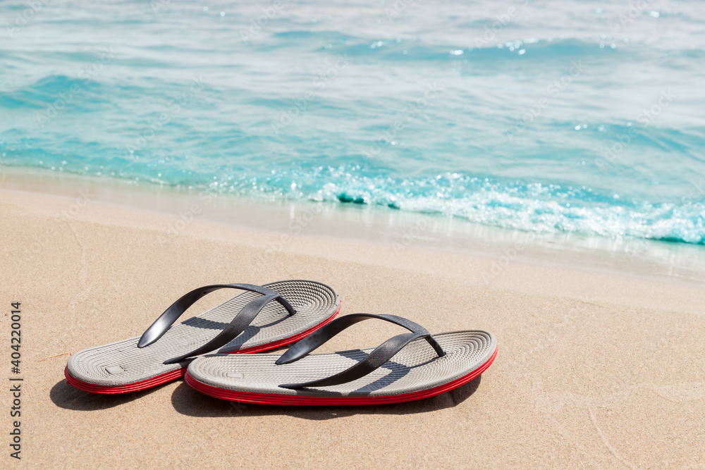 Obraz premium A pair of sandals with red soles on a sunny beach with sand near the seashore on a summer day. Rest and travel to hot countries. Vacation.