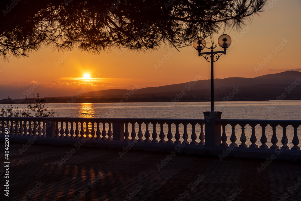 Summer Sunset in Golden tones. Silhouette of a balustrade. Shadow of the balustrade on the embankment. Back lighting. The sun sets in the mountains and is reflected in the sea. Pine branch in the