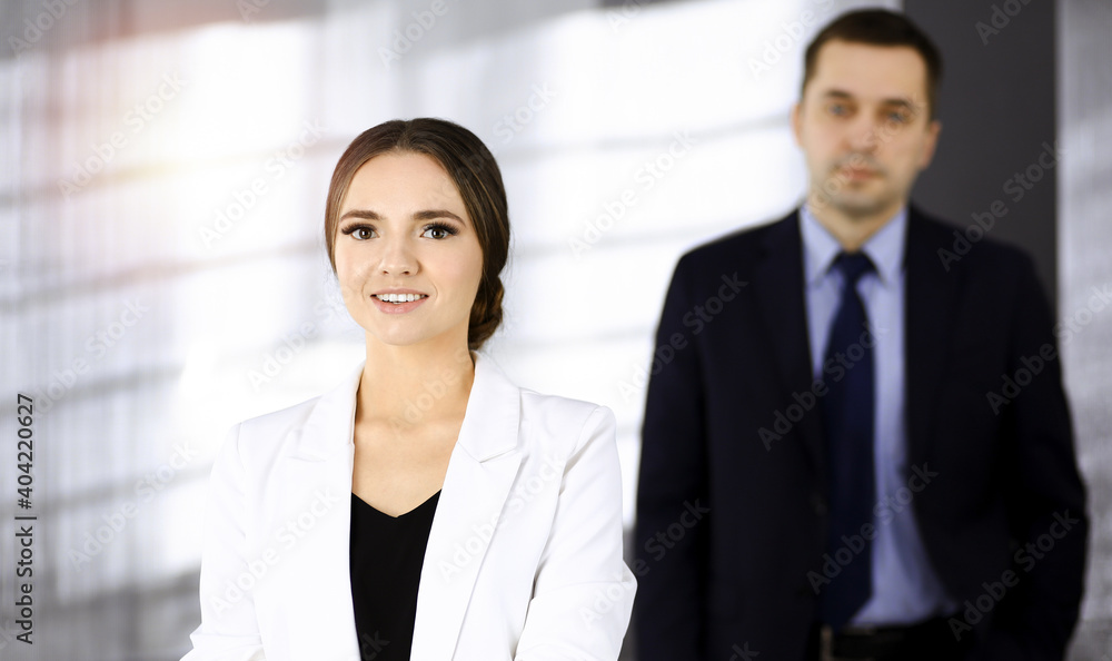 Young intelligent businesswoman is standing straight in a sunny modern office with her colleague at the background. Business people concept Young intelligent businesswoman is standing straight in a sunny modern office with her colleague at the background. Business people concept