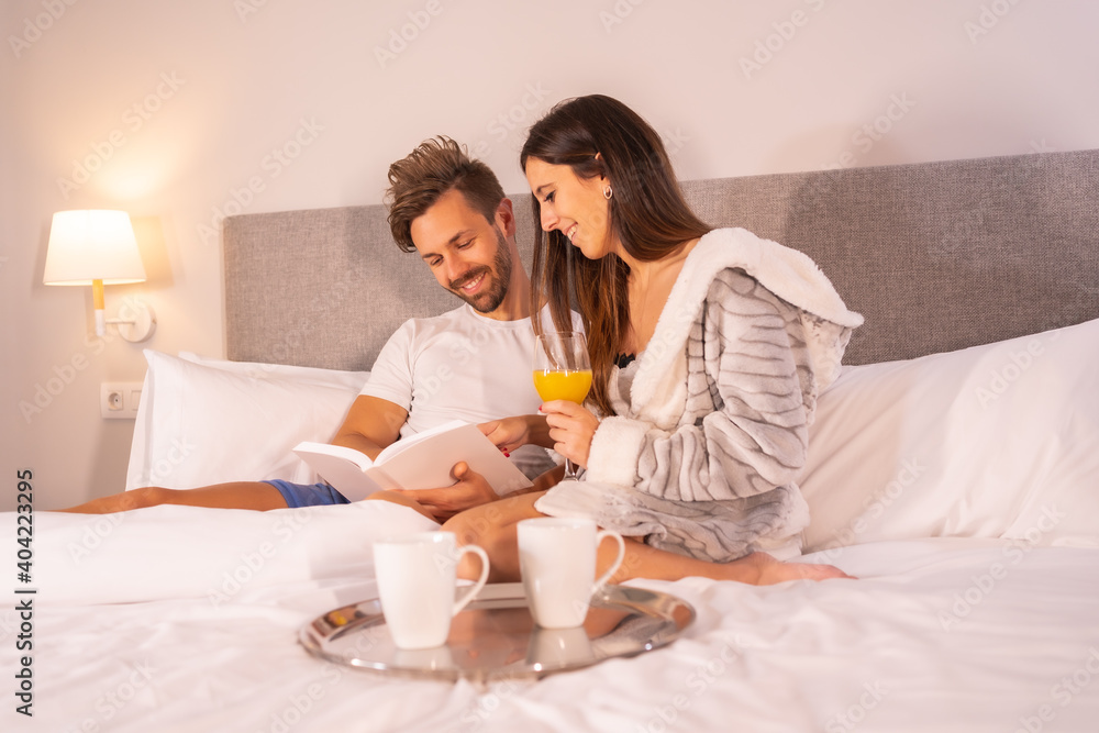 A couple in pajamas reading a book in the breakfast of coffee and orange juice in the hotel bed, lifestyle of a couple in love.