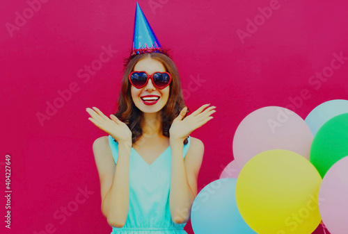 Happy surprised smiling young woman in birthday cap with colorful balloons on a pink background