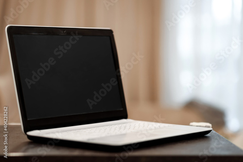 A laptop with a blank black screen in an angular position on a table against a blurred room background. The concept of workplace, office, coworking.