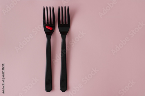 A red pill on a black fork. Two black forks with a red capsule on a pink background.