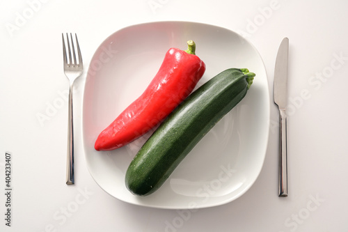 Raw red pointed pepper and zucchini on a white plate and cutlery on a bright background, healthy diet with mediterranean vegetables to lose weight, copy space, high angle view from above