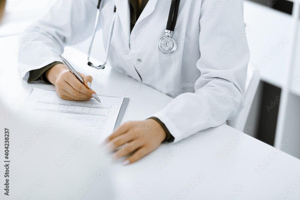 Unknown woman-doctor and female patient sitting and talking at medical examination in clinic, close-up. Therapist wearing green blouse is filling up medication history record. Medicine concept Unknown woman-doctor and female patient sitting and talking at medical examination in clinic, close-up. Therapist wearing green blouse is filling up medication history record. Medicine concept