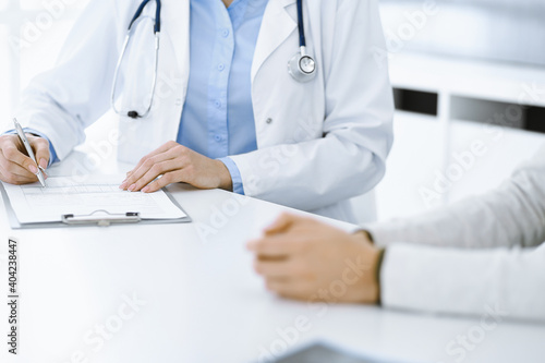 Unknown woman-doctor and female patient sitting and talking at medical examination in clinic  close-up. Therapist wearing blue blouse is filling up medication history record. Medicine concept