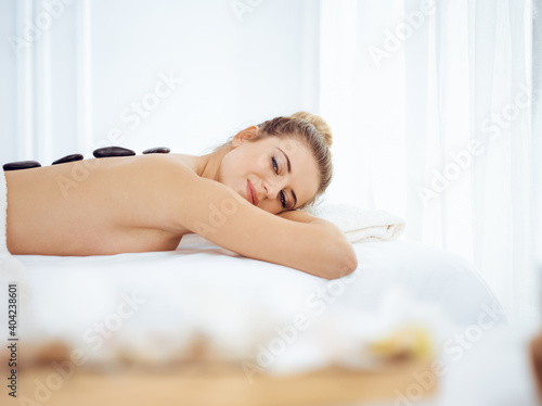 Young and blonde woman enjoying treatment with hot stones in spa salon. Beauty concept