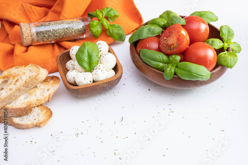 fresh tomatoes and green basil in a wooden bowl, near mozzarella, croutons and spices on a white table