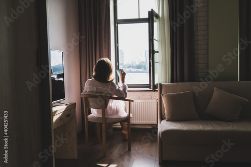 Back view of beautiful woman with cup of coffee in a transparent nightgown sitting on the armchair near open window.