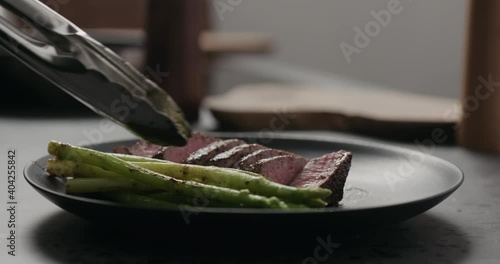 Slow motion orbit shot man serves sliced beef steak with asparagus on black plate on concrete countertop photo