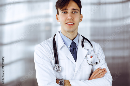 Cheerful smiling man-doctor standing with arms crossed in clinic. Perfect medical service with young smart physician in hospital. Medicine concept