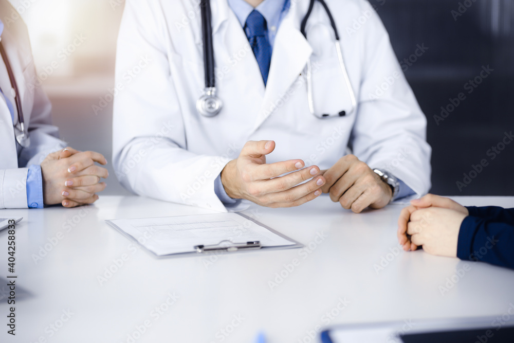 Two doctors and patient woman discussing something while sitting at the desk in modern clinic. Perfect medical service, medicine concept Two doctors and patient woman discussing something while sitting at the desk in modern clinic. Perfect medical service, medicine concept