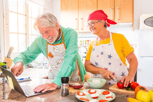 couple of two happy seniors cooking together in the kitchen learning and following a video tutorial of cook in the laptop -  preparing healthy food together photo