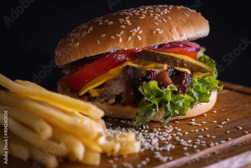 burger with baked slices of fried meat, tomatoes, onions, cheese, lettuce and french fries on a wooden surface isolated on a dark background. The concept of delicious food