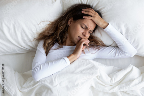 Top view unhappy woman with closed eyes touching forehead, suffering from headache, lying under blanket in bed alone, frustrated young female feeling unhealthy, strong sudden pain, hangover
