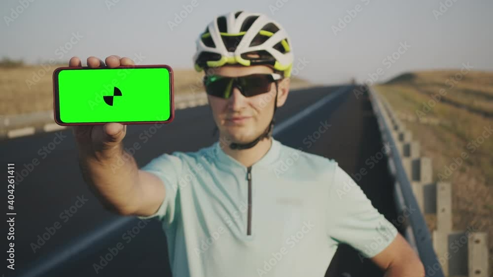 The cyclist shows the camera a horizontal smartphone with a green screen with marker on the background of the highway outdoors. A young man in a helmet and sunglasses holds the phone at arm's length The cyclist shows the camera a horizontal smartphone with a green screen with marker on the background of the highway outdoors. A young man in a helmet and sunglasses holds the phone at arm's length