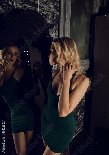 Sexy fit blond model with curly hair style in  fashion dreen dress looking on her reflection in the mirror on dark dramatic Gothic background in dark shadow light. Mystic atmosphere. Stunning