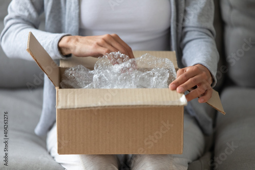 Close up woman opening cardboard box with fragile goods or sending package, sitting on couch at home, young female client customer received online store order, quick delivery service concept